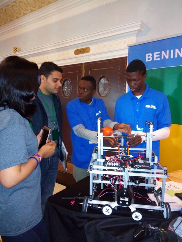Des membres de la Team Benin en polo bleu échangent avec des visiteurs autour de leur robot métallique posé sur une table, devant une bannière aux couleurs du Bénin