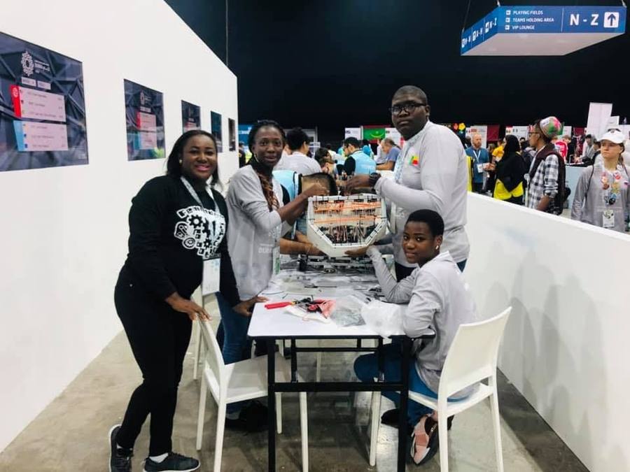 Une mentor et des membres de la Team Benin en noir et gris posent autour d'une table de travail sur laquelle est posé un robot, dans un grand hall d'exposition
