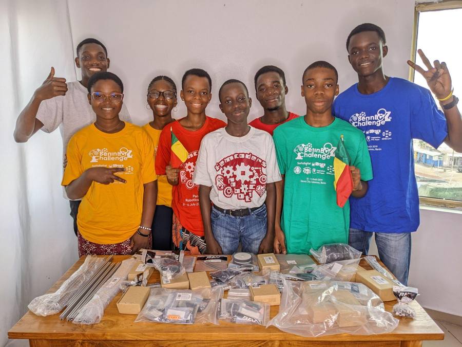 Un groupe de jeunes en t-shirts de couleur pose en souriant derrière une table couverte de sachets de pièces et de profilés métalliques, un drapeau béninois à la main