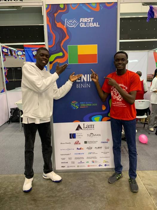 Deux jeunes de la Team Benin, l'un en sweat blanc et l'autre en t-shirt rouge, désignent en souriant le drapeau du Bénin sur le panneau officiel de leur stand FIRST Global