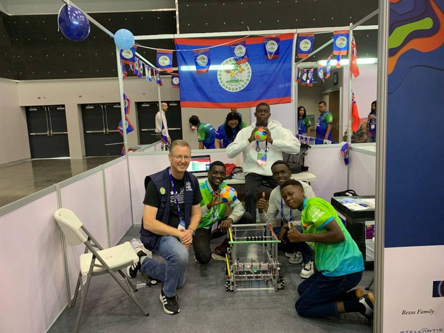 Des membres de la Team Benin accroupis dans leur stand de compétition autour d'un robot métallique, avec un mentor souriant